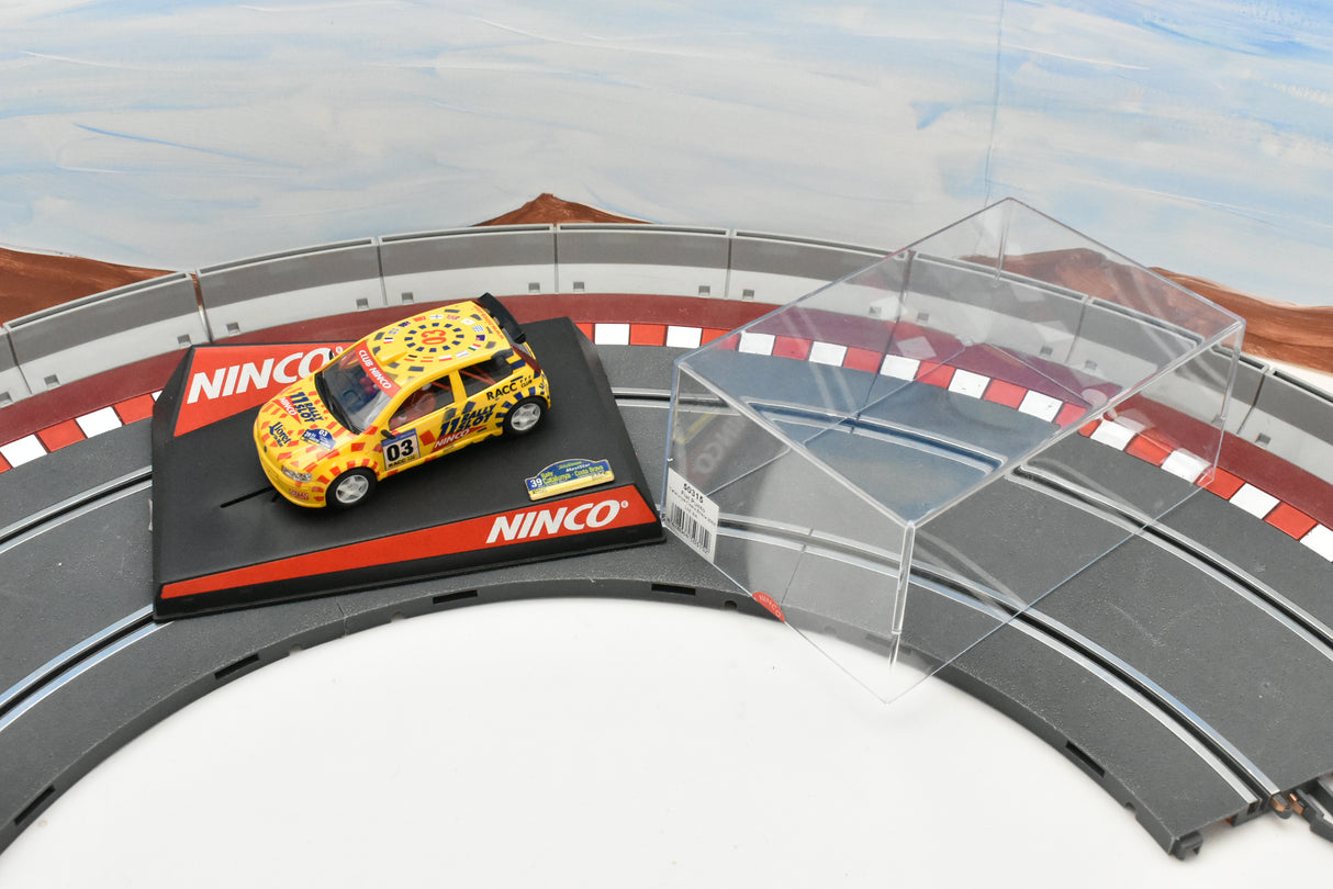 Yellow race car on a track with a clear barrier, set against a scenic backdrop for Ninco 50315
