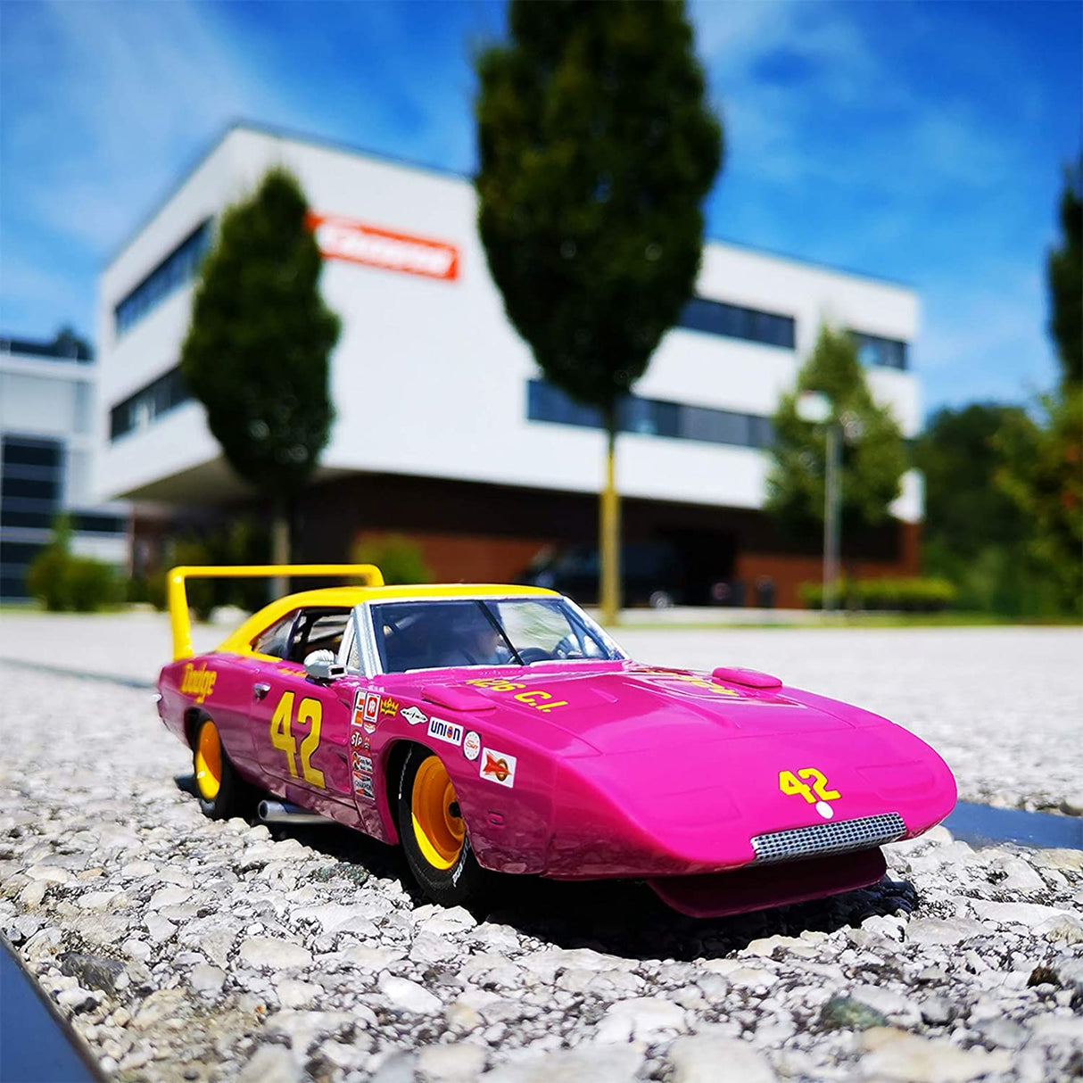 Pink Dodge Charger Daytona race car on gravel with modern building backdrop for Carrera Evolution 20027638