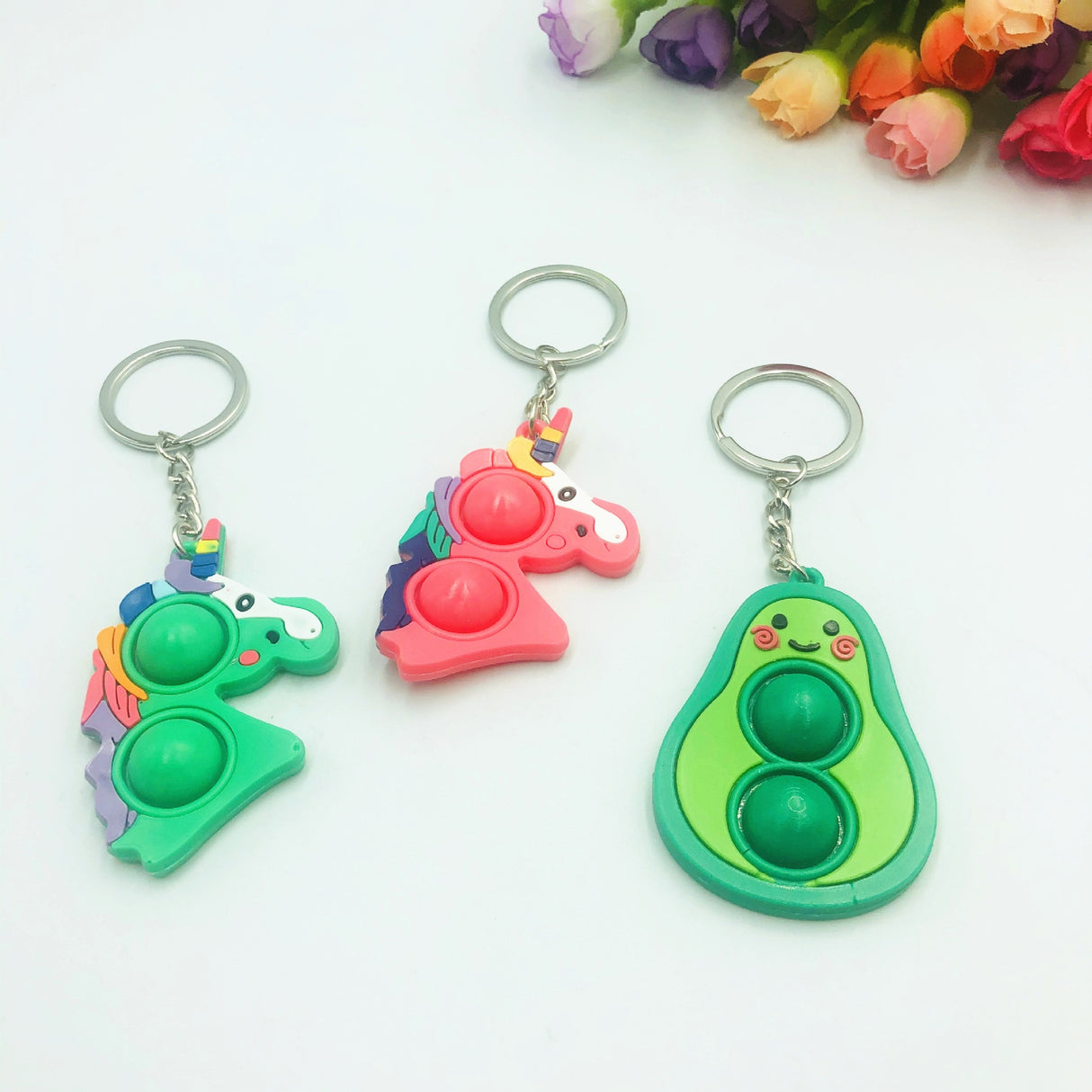 Three colorful, pop-it style keychain fidget toys: a green dinosaur, a pink unicorn, and a green avocado for Faire FH2928