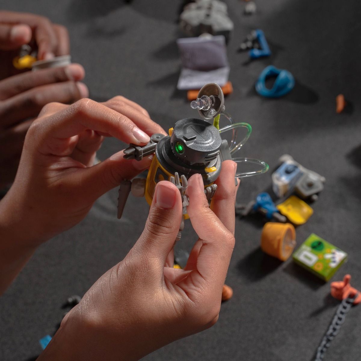 Hands assembling a small, mechanical robot toy among assorted junk parts for HexBug 430-6843, parts view, product view 8