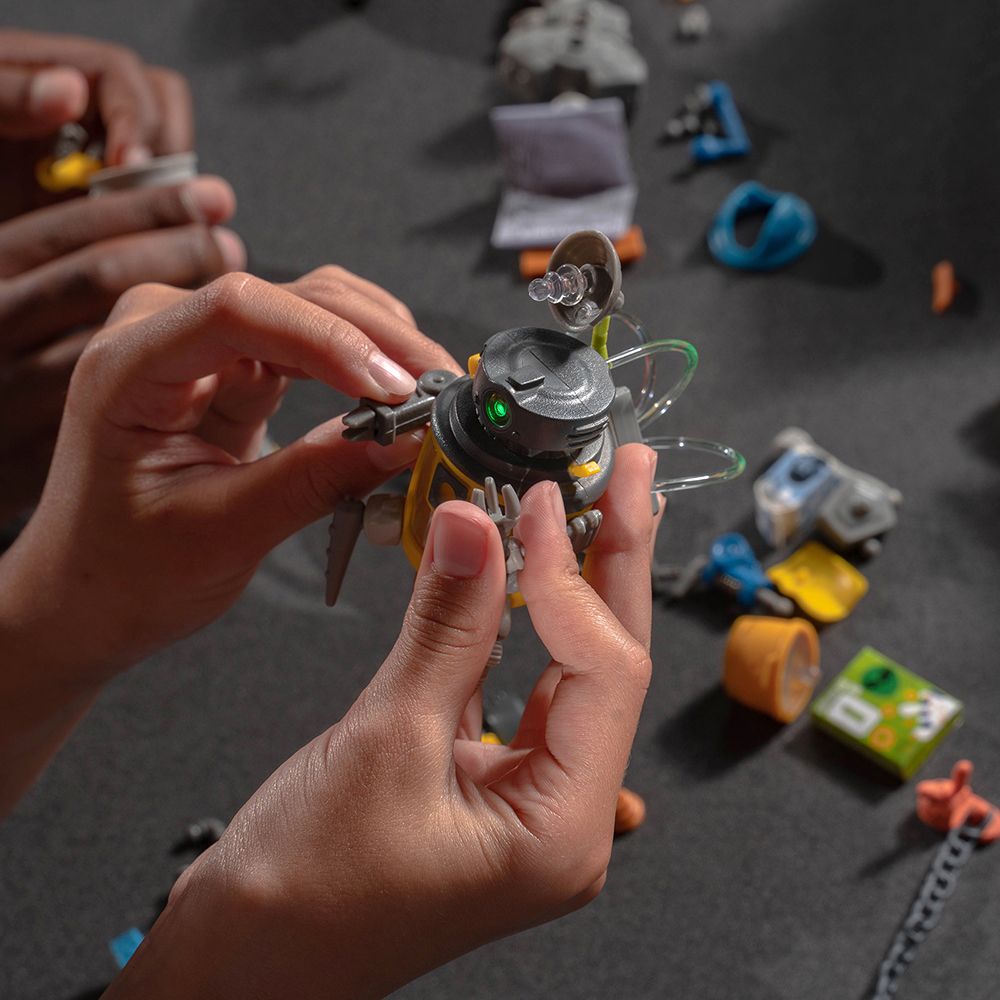 Hands assembling a small, mechanical robot toy among assorted junk parts for HexBug 430-6843, parts view, product view 13