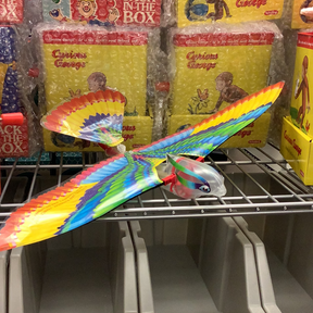 Colorful toy bird toy resting on a metal rack near yellow boxes for Schylling NTN